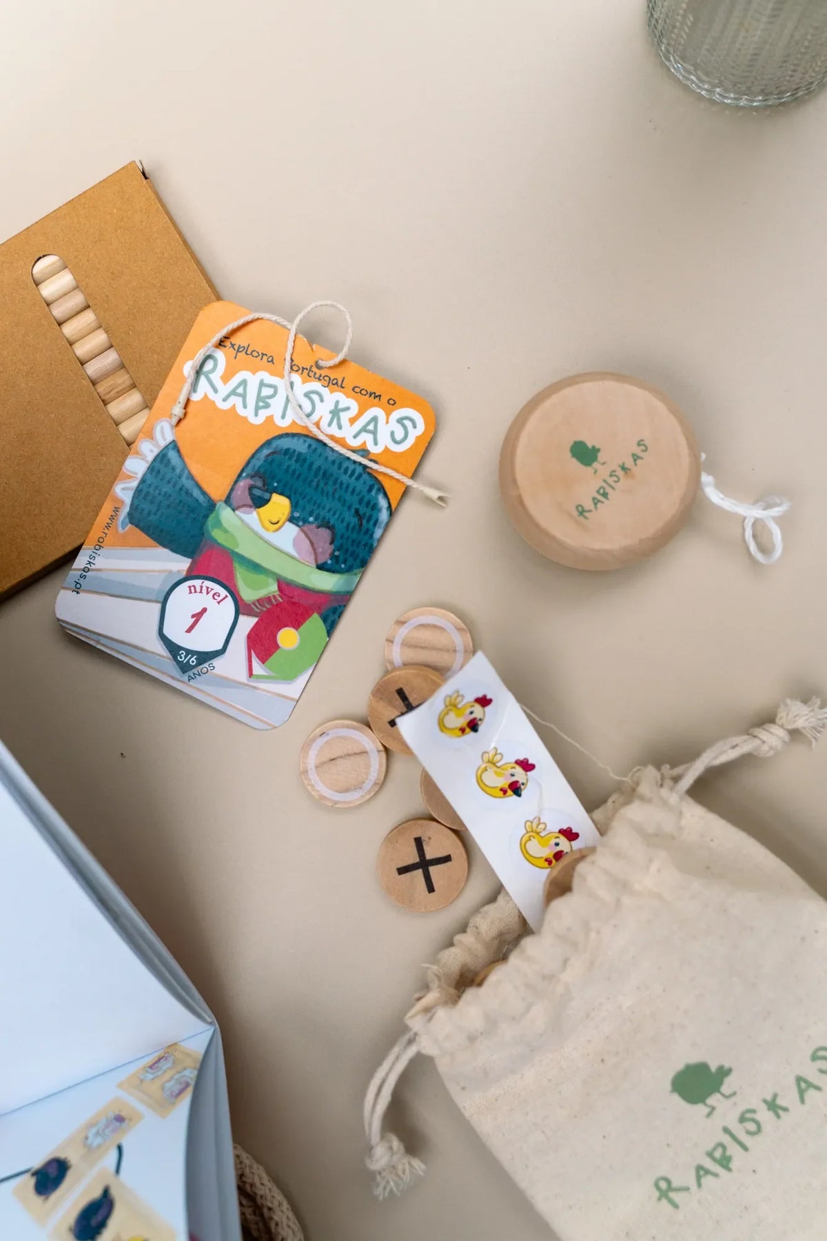 Conteúdo do Kit Rabiskas Nível 1: caderno de atividades, io-iô de madeira, jogo do galo, lápis de cor e saco Rabiskas